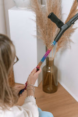 Person holding a colourful leopard crutch with decorative elements in a minimalistic indoor setting.